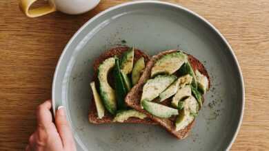 Overhead view of unrecognizable person holding big round plate with sandwich with avocado near cup of tea on light brown wooden table in bright room