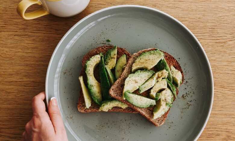 Overhead view of unrecognizable person holding big round plate with sandwich with avocado near cup of tea on light brown wooden table in bright room