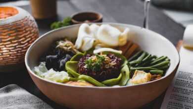 From above of appetizing plate with green beans and batata pieces and different types cabbage and squid pieces with avocado slices and red rice in middle of oriental dish served with glass of drink on table of restaurant