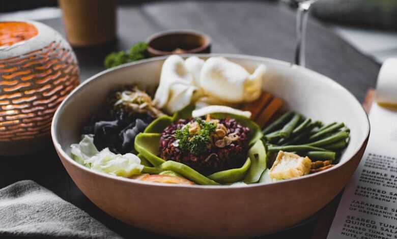 From above of appetizing plate with green beans and batata pieces and different types cabbage and squid pieces with avocado slices and red rice in middle of oriental dish served with glass of drink on table of restaurant