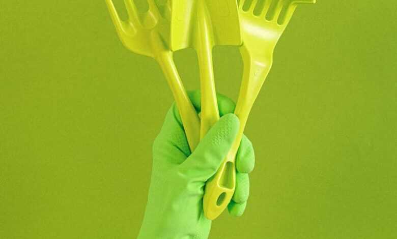 Crop anonymous person wearing rubber glove showing set of gardening tools including trowel and hand rake against green background